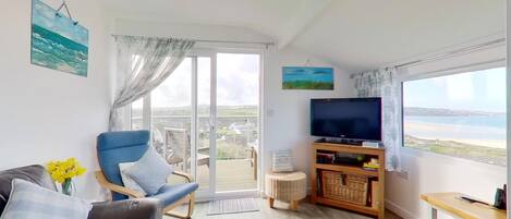 Un salon comprenant un canapé, un fauteuil, un téléviseur posé sur un meuble en bois, et une vue sur la mer grâce à de grandes fenêtres.