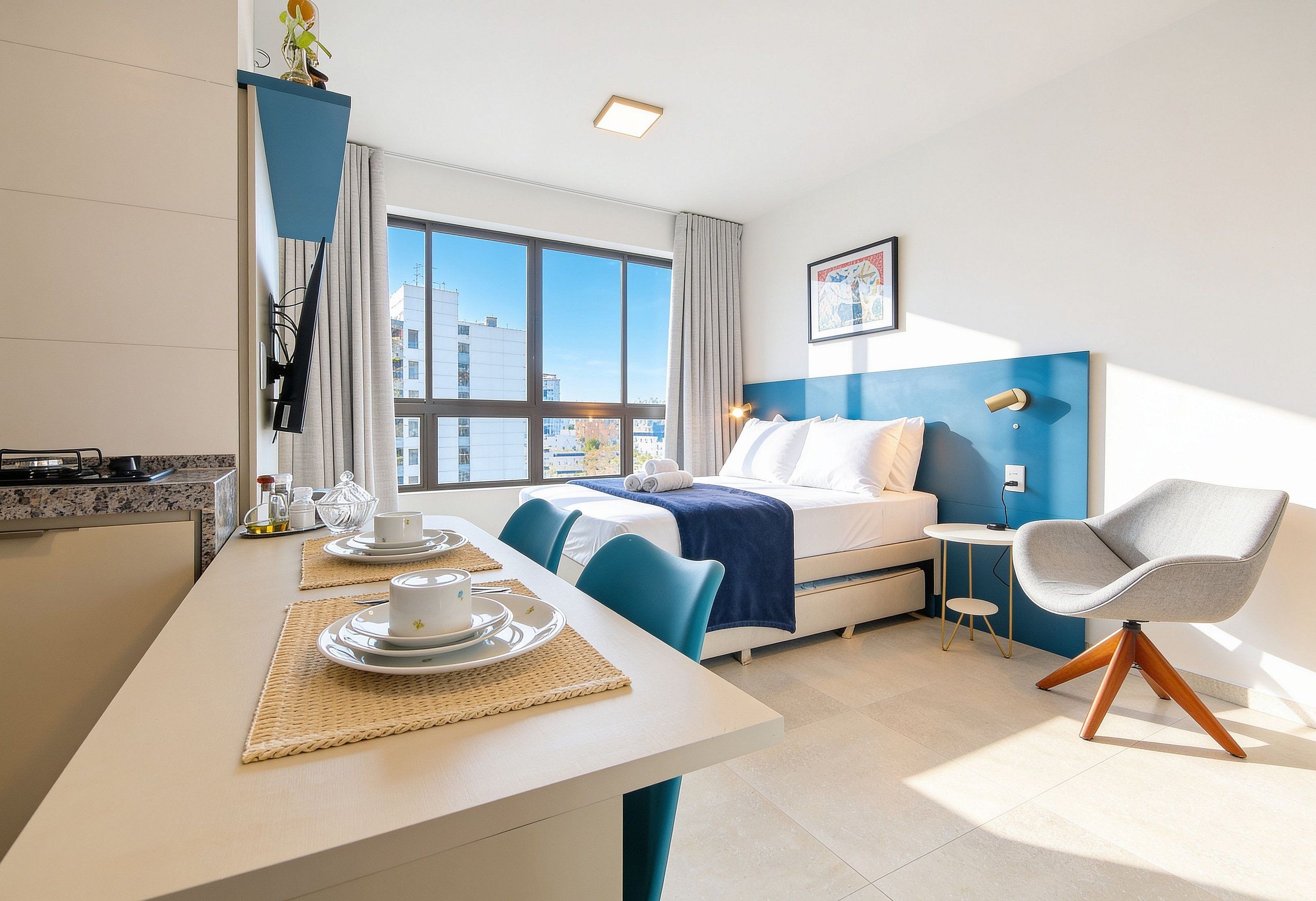 Quarto de hotel com uma cama, uma mesa de trabalho com cadeira, uma mesa de jantar com cadeira e vista da cidade através de amplas janelas.