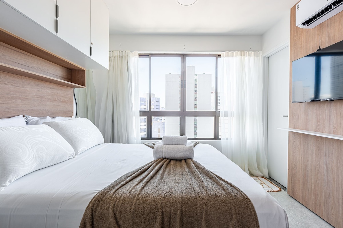 Una habitación de hotel con cama, televisión, ventana con cortinas y vistas a edificios.