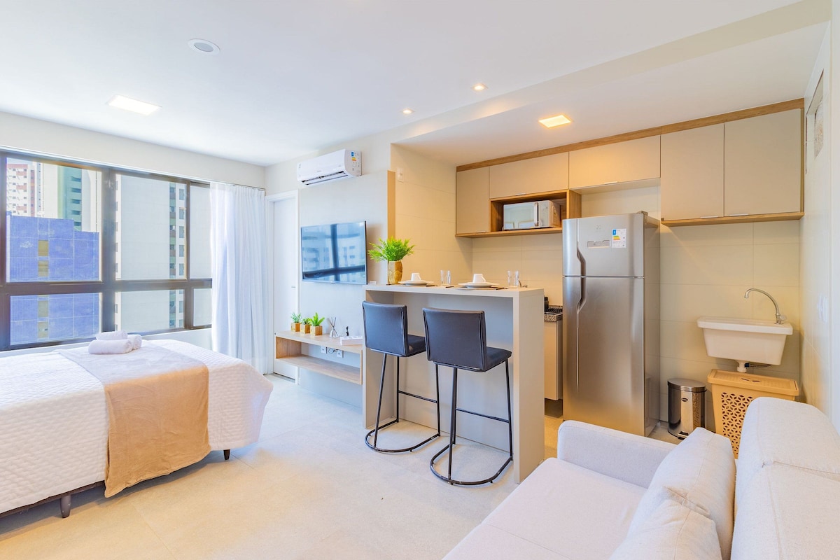 A modern hotel room with a large bed, a small kitchenette, a sitting area with a sofa, and a large window offering a city view.
