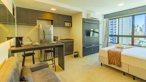 A compact hotel room with a kitchenette, a sofa, a bed, and a large window offering a city view.