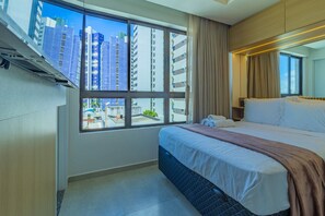 Ein Hotelzimmer mit großem Fenster, einem Bett, einem Nachttisch und Blick auf hohe Gebäude.