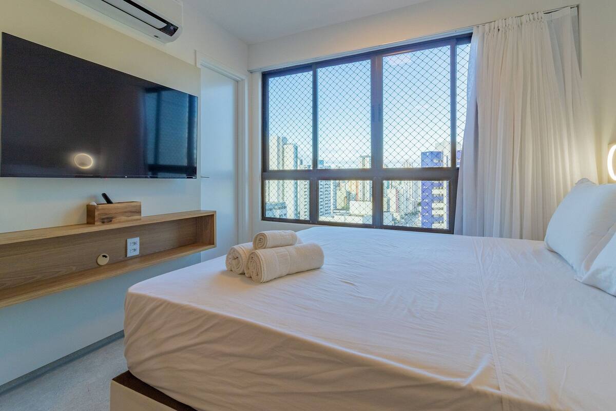 Habitación de hotel con una cama grande, un televisor montado en la pared, una ventana con vistas a la ciudad y un estante de madera con un objeto pequeño.