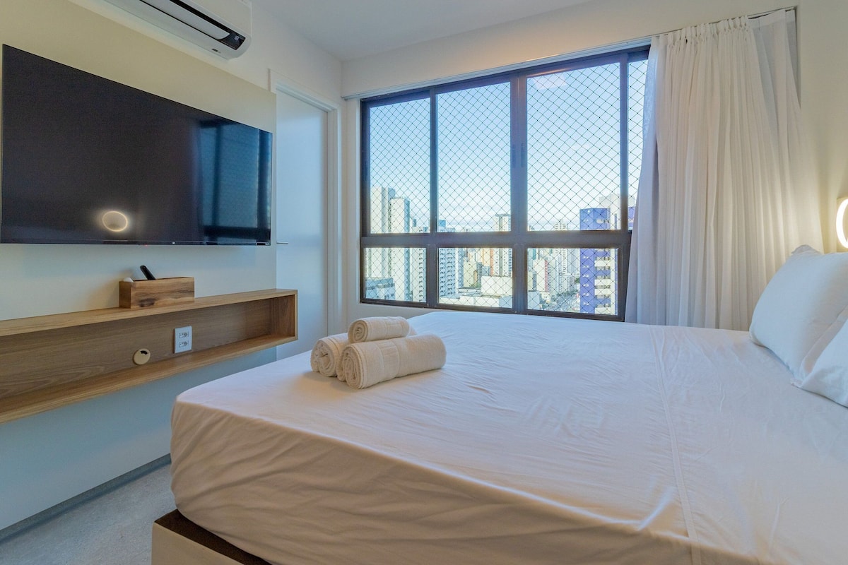A hotel room with a large bed, a TV mounted on the wall, a window with a city view, and a wooden shelf with a small item on it.