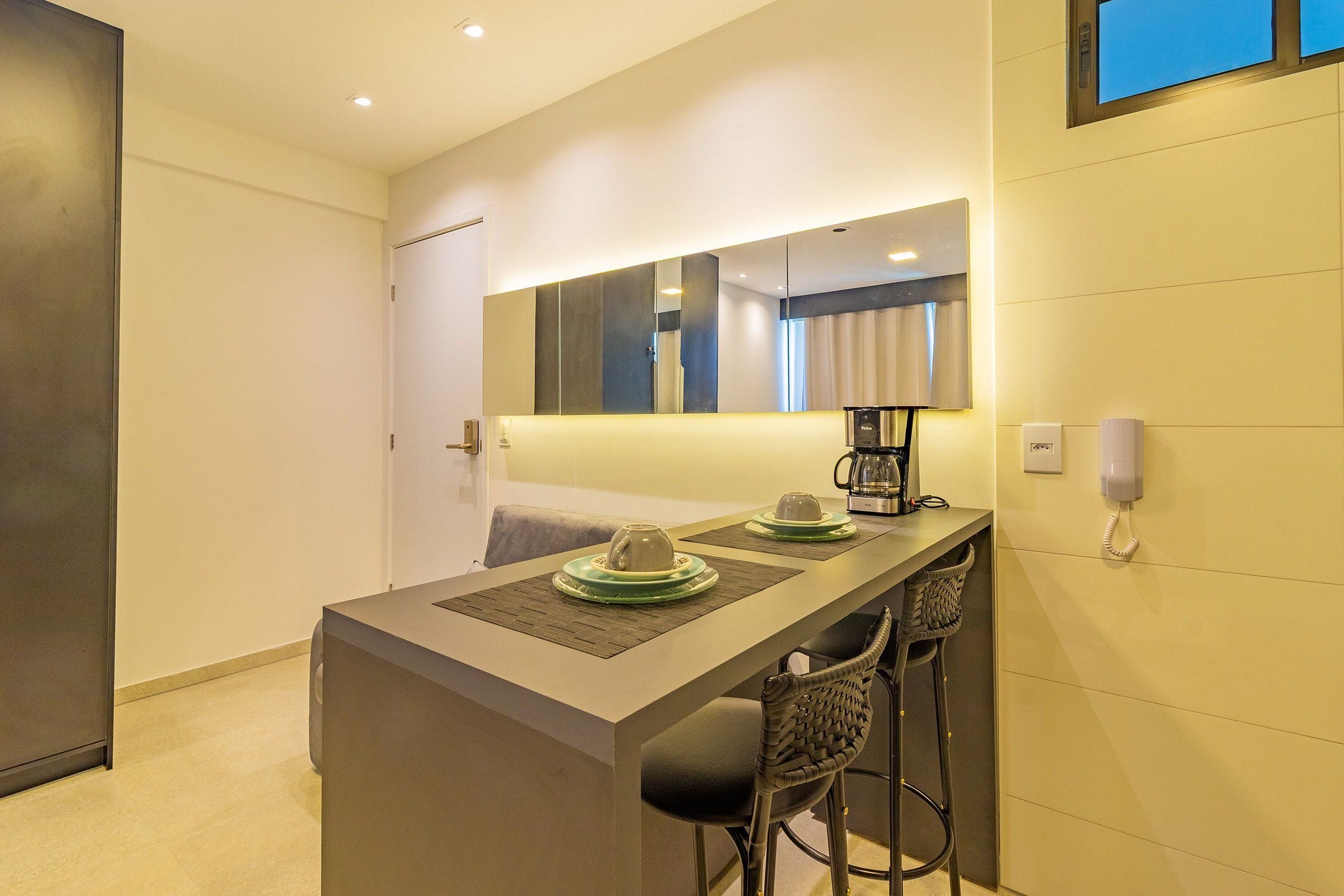 Quarto de hotel moderno com uma mesa de jantar comprida, duas cadeiras, uma máquina de café e um telefone na parede.