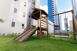 Un'area giochi con uno scivolo e altalene, circondata da un prato e con alti edifici sullo sfondo.