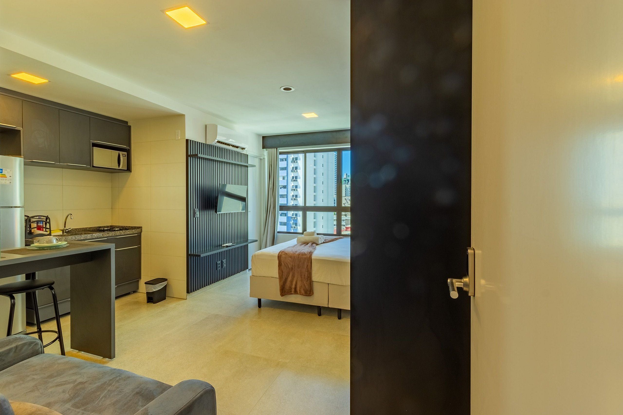 Cozinha moderna com micro-ondas, uma pequena ilha e um banquinho de bar. Há uma cama com um cobertor marrom e uma janela com vista para edifícios.