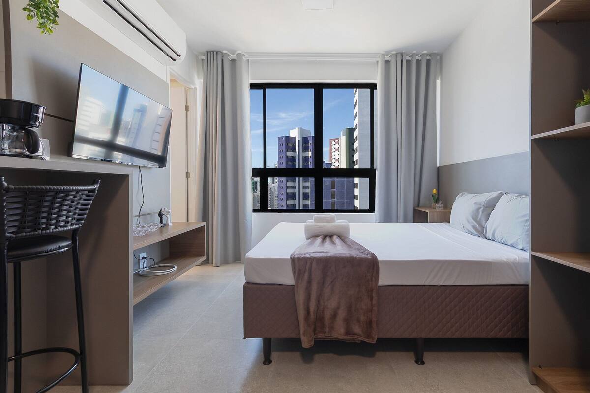 Habitación de hotel moderna con cama, televisor, escritorio y cafetera, y ventana con vista a la ciudad.