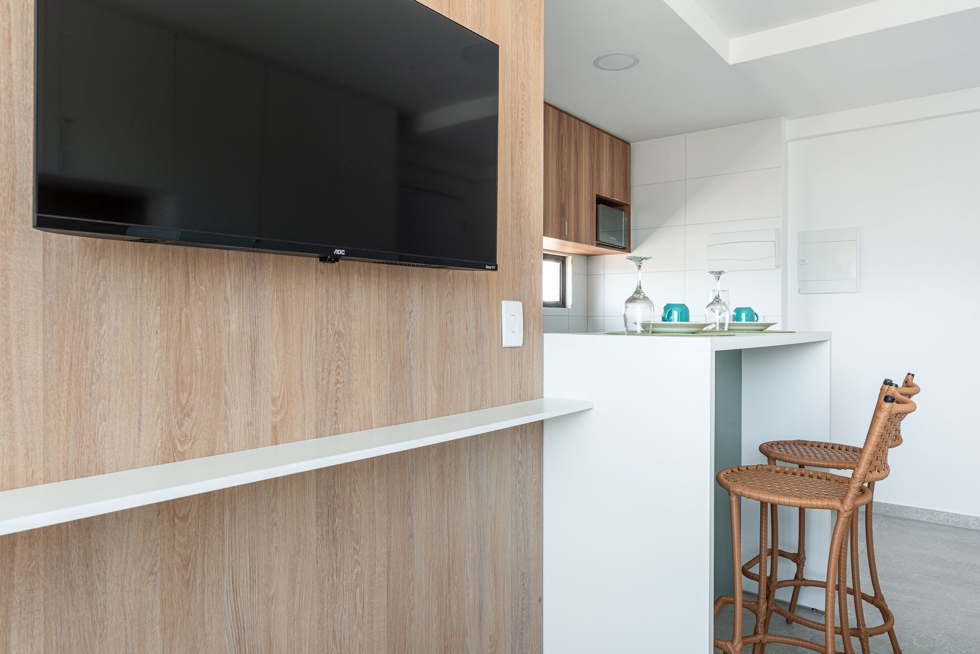 Una cocina moderna con un televisor de pantalla plana montado en una pared de madera, una encimera blanca y taburetes de bar de mimbre.