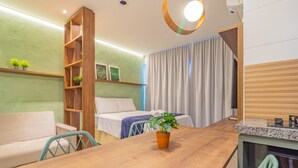A modern hotel room with a wooden dining table, a sofa, a bed with a blue blanket, a wooden shelf with plants, and a large window with curtains.
