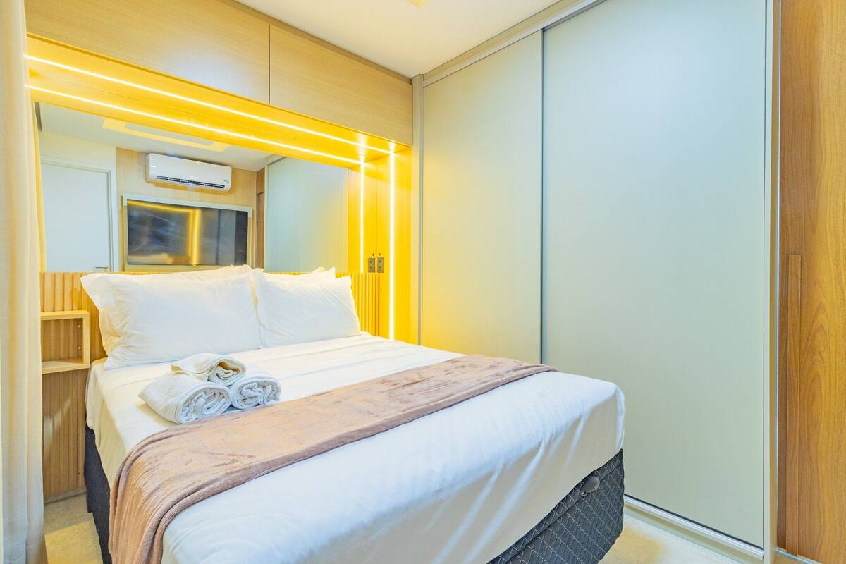 Una habitación de hotel moderna con una cama bien hecha, una mesita de noche, un televisor integrado y un espejo grande.