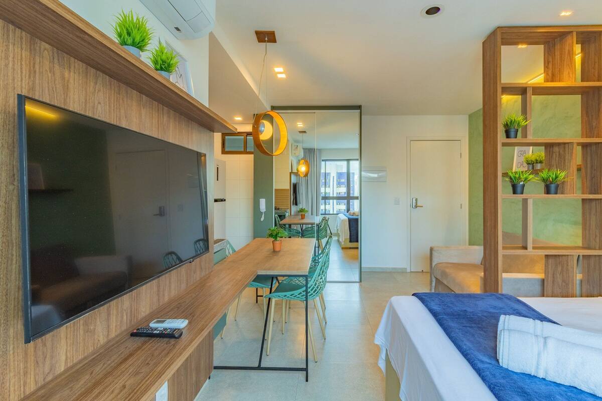 Una habitación de hotel moderna con un televisor de pantalla plana grande, un escritorio de madera con un control remoto, una zona de comedor con sillas, una cama con una colcha azul y un estante de madera con plantas.