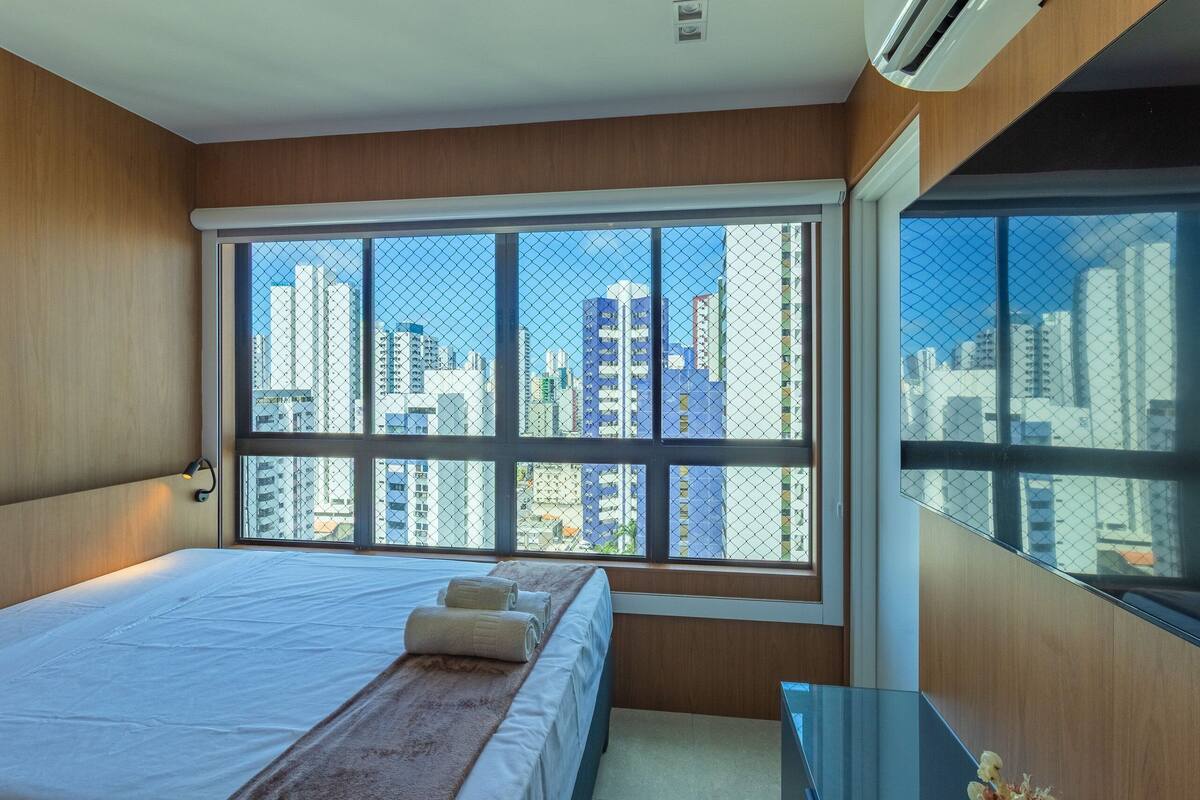 Habitación de hotel moderna con un amplio ventanal que ofrece vistas a la ciudad, una cama bien hecha y una mesita.