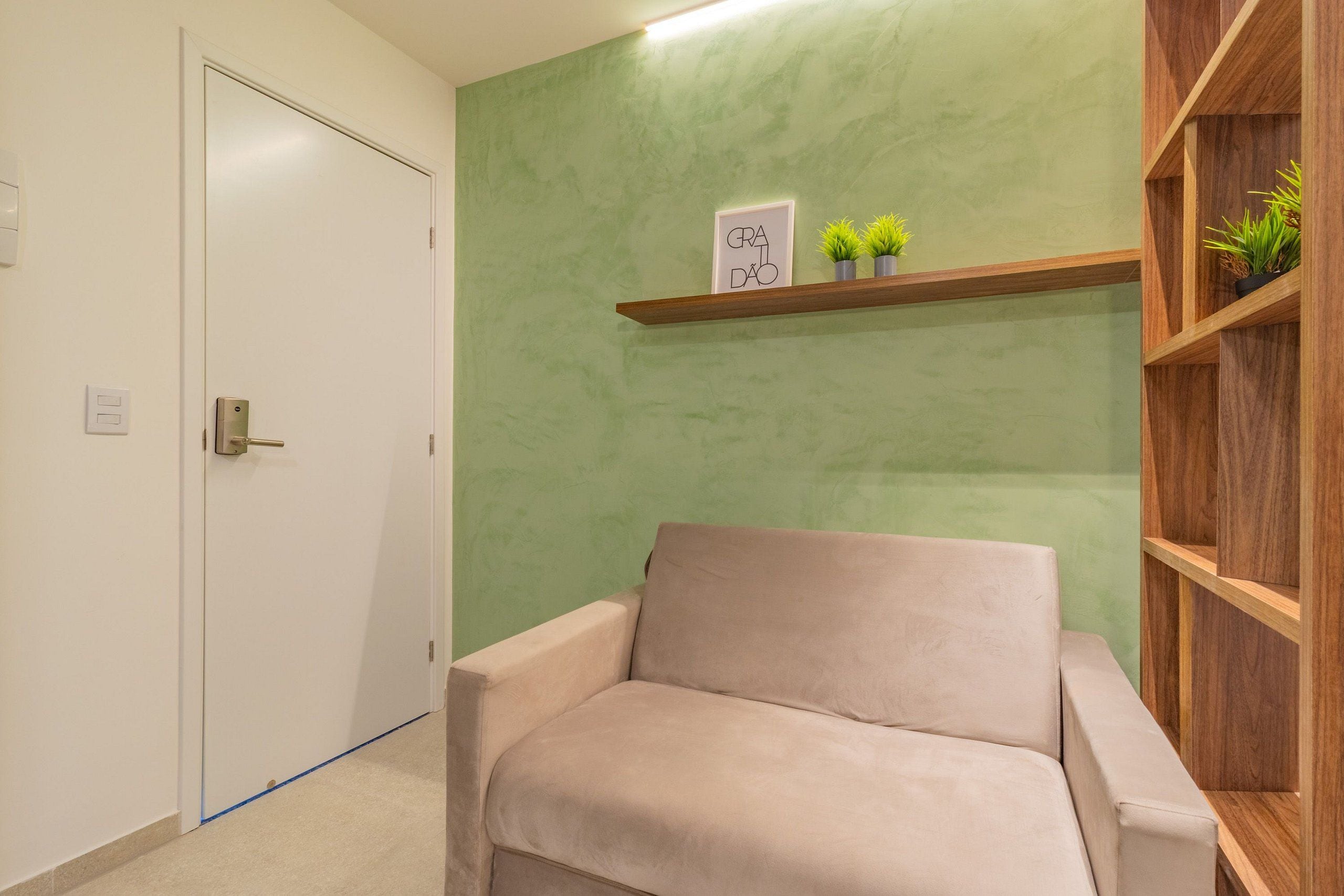 A beige armchair in a room with a green textured wall, a wooden shelf with plants, and a white door.