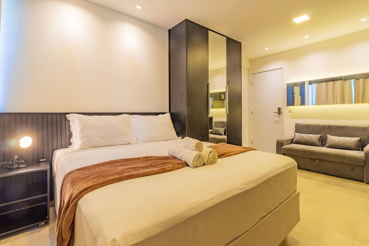 Una habitación de hotel moderna con una cama grande, una mesita de noche con lámpara, un armario, un sofá y un espejo.