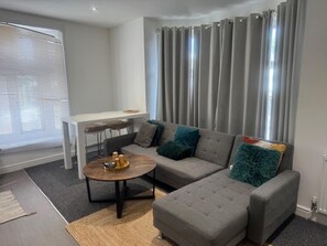 A modern living room with a grey sectional sofa, a round coffee table, and a white desk with two chairs. - Lewisham - Apartment - Sleeps 5 - Central Location (London)
