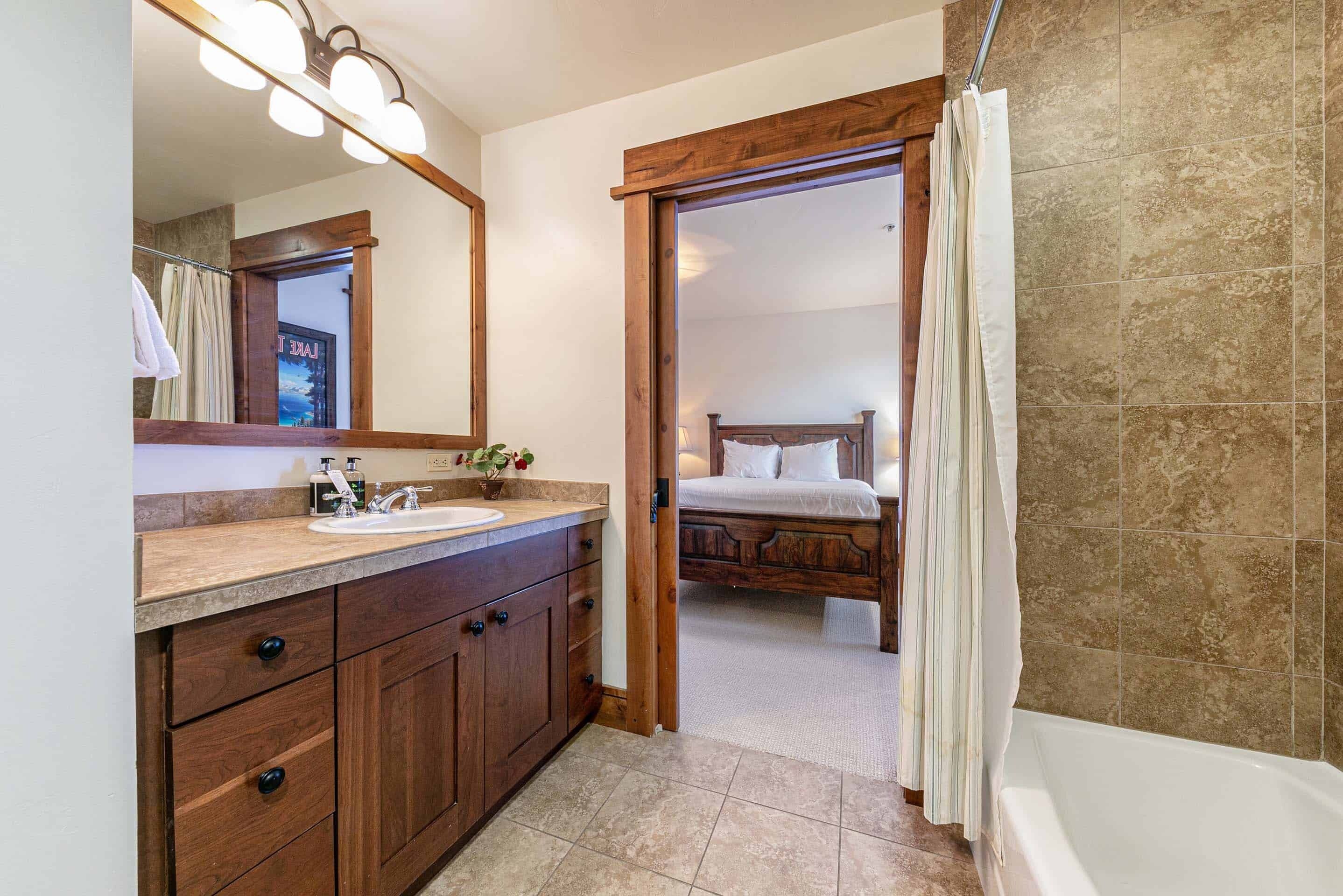 A bathroom with a wooden vanity, a large mirror, a bathtub, and a wooden bed in the adjacent room.