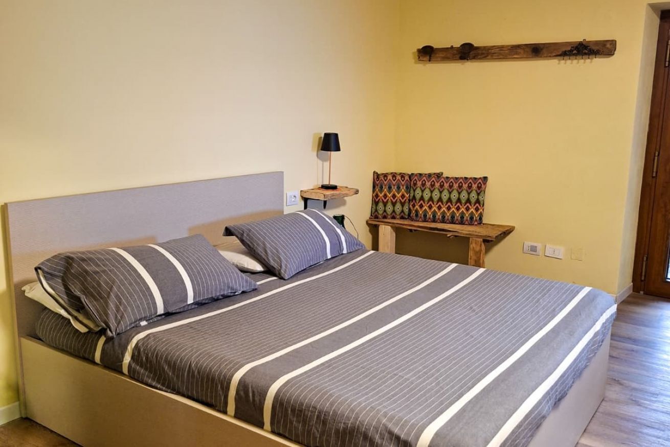 A bed with striped bedding, a bedside table with a lamp, a colorful cushion, and a wooden shelf on the wall.