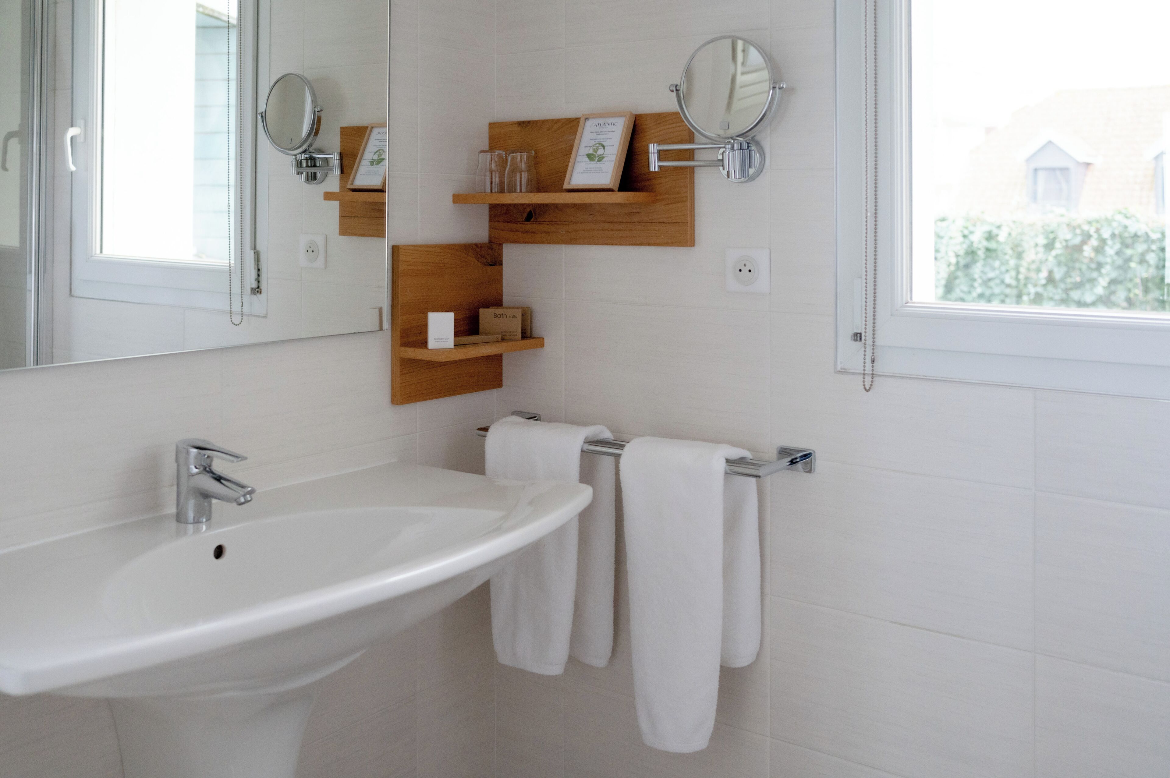 Un baño con un lavamanos blanco, dos espejos, repisas de madera y una ventana.
