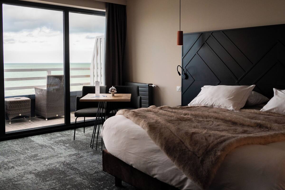 Une chambre d’hôtel avec un lit, un bureau, une chaise, une vue sur l’océan et une grande fenêtre.