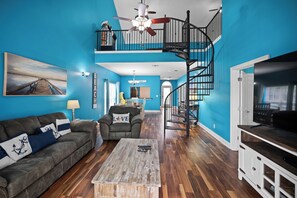 A living room with a blue wall, a wooden coffee table, a sofa, a TV stand, and a staircase. - 🏝️Gated Community-Great Amenities-Coral Retreat🌊 (Pensacola)