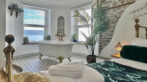 Ein Schlafzimmer mit einer großen Badewanne, einem Bett mit einer grün-weißen Bettdecke und Meerblick durch das Fenster.