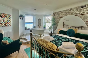 Un dormitorio con pared de piedra, una cama con ropa de cama verde y blanca, una bañera y vista al mar a través de una ventana.
