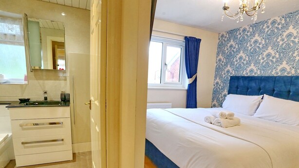 A bedroom with a bed, a window with blue curtains, a blue headboard, and a bathroom with a mirror and sink.