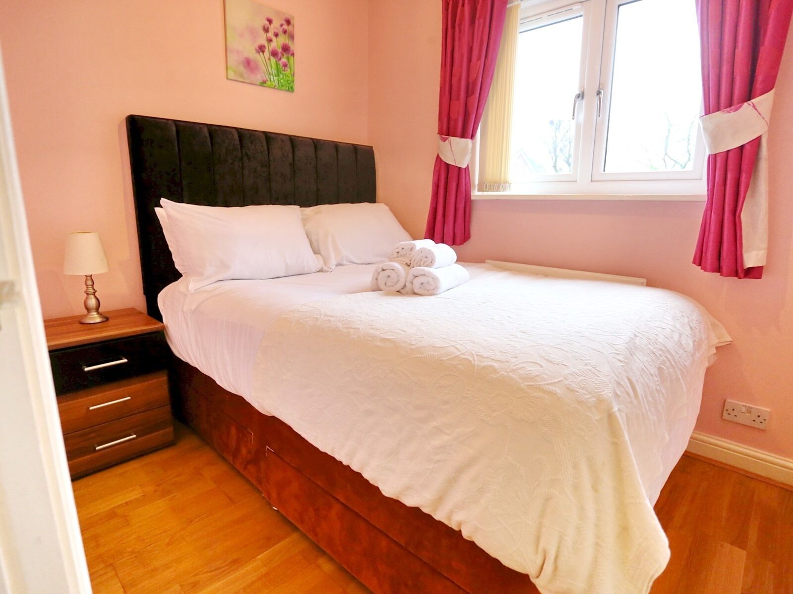 Una cama bien hecha en una habitación con piso de madera, una mesita de noche con una lámpara y una ventana con cortinas.