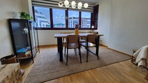 Un espace repas comprenant une table, des chaises, un tapis, un meuble et une fenêtre donnant sur des immeubles.