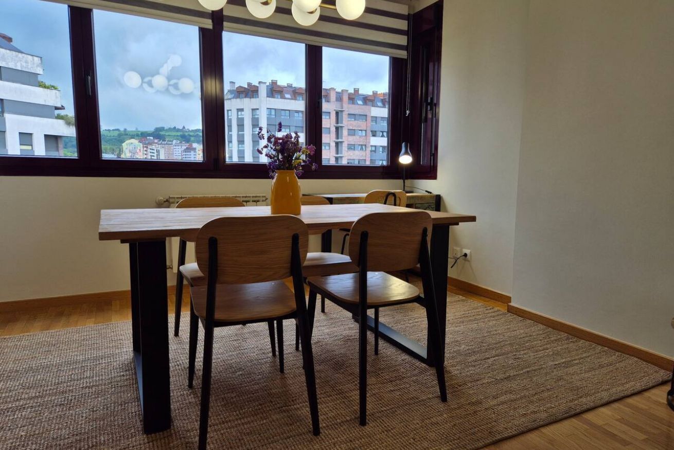 Un comedor con mesa y sillas, un jarrón con flores, y vistas a edificios a través de amplios ventanales.