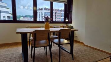 Un comedor con mesa y sillas, un jarrón con flores, y vistas a edificios a través de amplios ventanales.