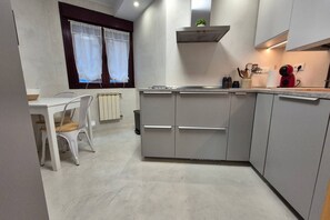 Cocina compacta con muebles blancos, horno empotrado y una ventana con cortinas traslúcidas.