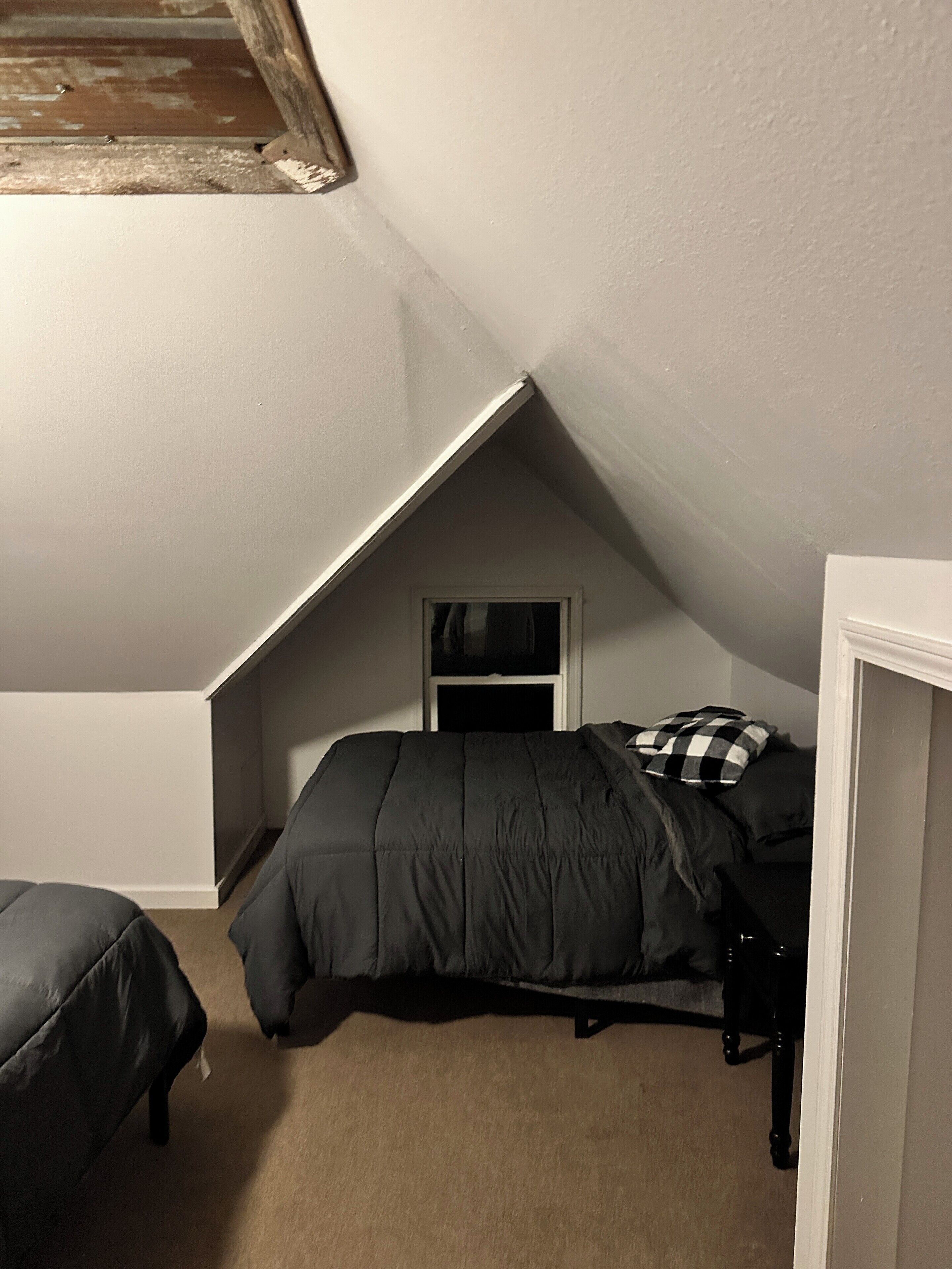 A bedroom with a sloped ceiling, a bed with a dark-colored comforter, a small window, and a door.