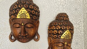 To træbuddhahoveder med detaljerede udskæringer og guldornamenter, monteret på en væg med tapet i et mønstret design.