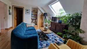 En hyggelig loftrum-stue med blå sofa, gule lænestole, en reol, et fjernsyn på et træskab og et ovenlysvindue.