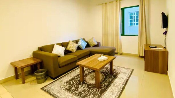 A living room with a sofa, two coffee tables, a patterned rug, a small side table, a waste basket, and a window with curtains. - Rest-Inn Two Bedrooms Apartment (Muscat)