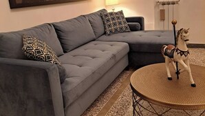 A living room with a grey sectional sofa, patterned throw pillows, a round coffee table with a decorative horse figurine, a large abstract painting, and a framed portrait on the wall.