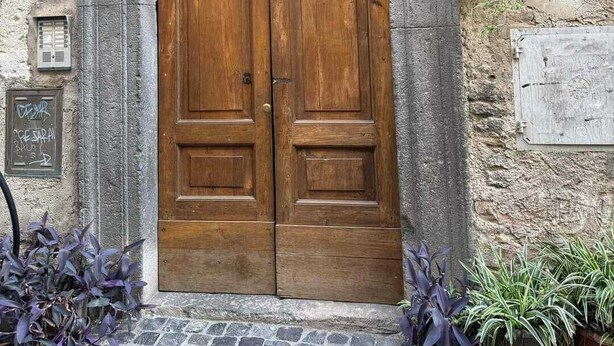 A stone building with a wooden double door, a small window above the door, and a potted plant hanging on the wall.