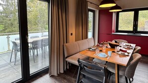A dining area with a wooden table set for four, a sofa, and a view of a body of water through large windows. - Modernes Chalet in Hahnenklee (Goslar)