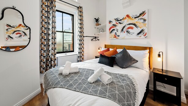 A neatly made bed with a geometric patterned bedspread, a bedside table with a lamp, and abstract paintings on the walls. - The Hallmark 3BR Apt JQ Birmingham, Parking & WiFi (Birmingham)