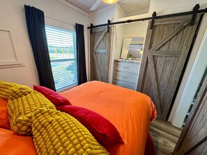 A bedroom with a bed, a wooden sliding door, and a window with blinds. - The Tiny Tejas with Pool & Hot Tub (Austin)