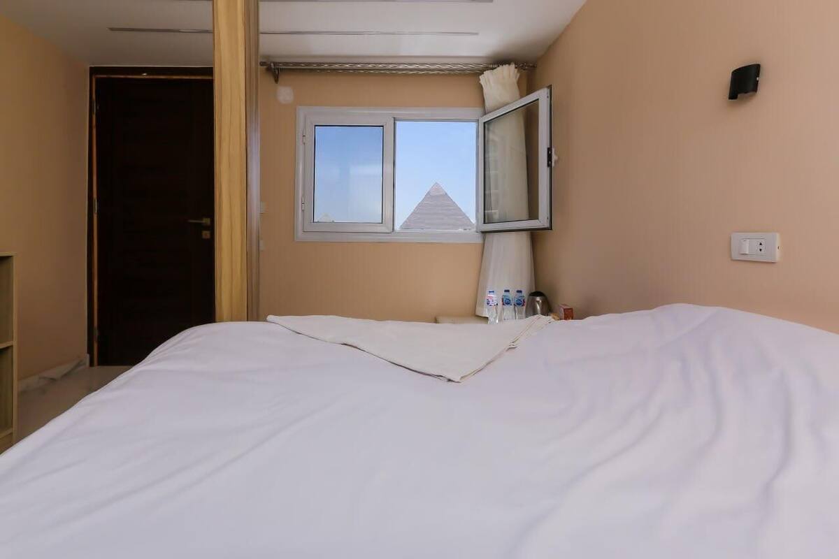A hotel room with a bed, a window with a view of a building, a bedside table with water bottles, and a power outlet.