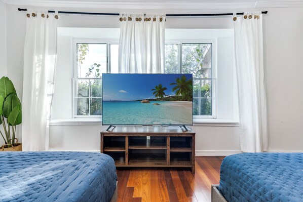 Ein Schlafzimmer mit Fernseher auf einem Holzständer, zwei Betten, einem Fenster mit Vorhängen und einer Topfpflanze.