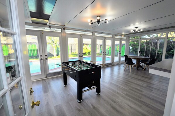 A spacious indoor area with a foosball table, a dining table with chairs, and large windows providing views of a backyard with a pool.