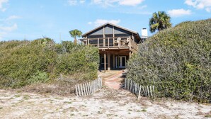 Une maison en bois à deux étages, dotée d’un balcon, entourée d’une végétation dense et bordée d’un chemin de sable menant à l’entrée.