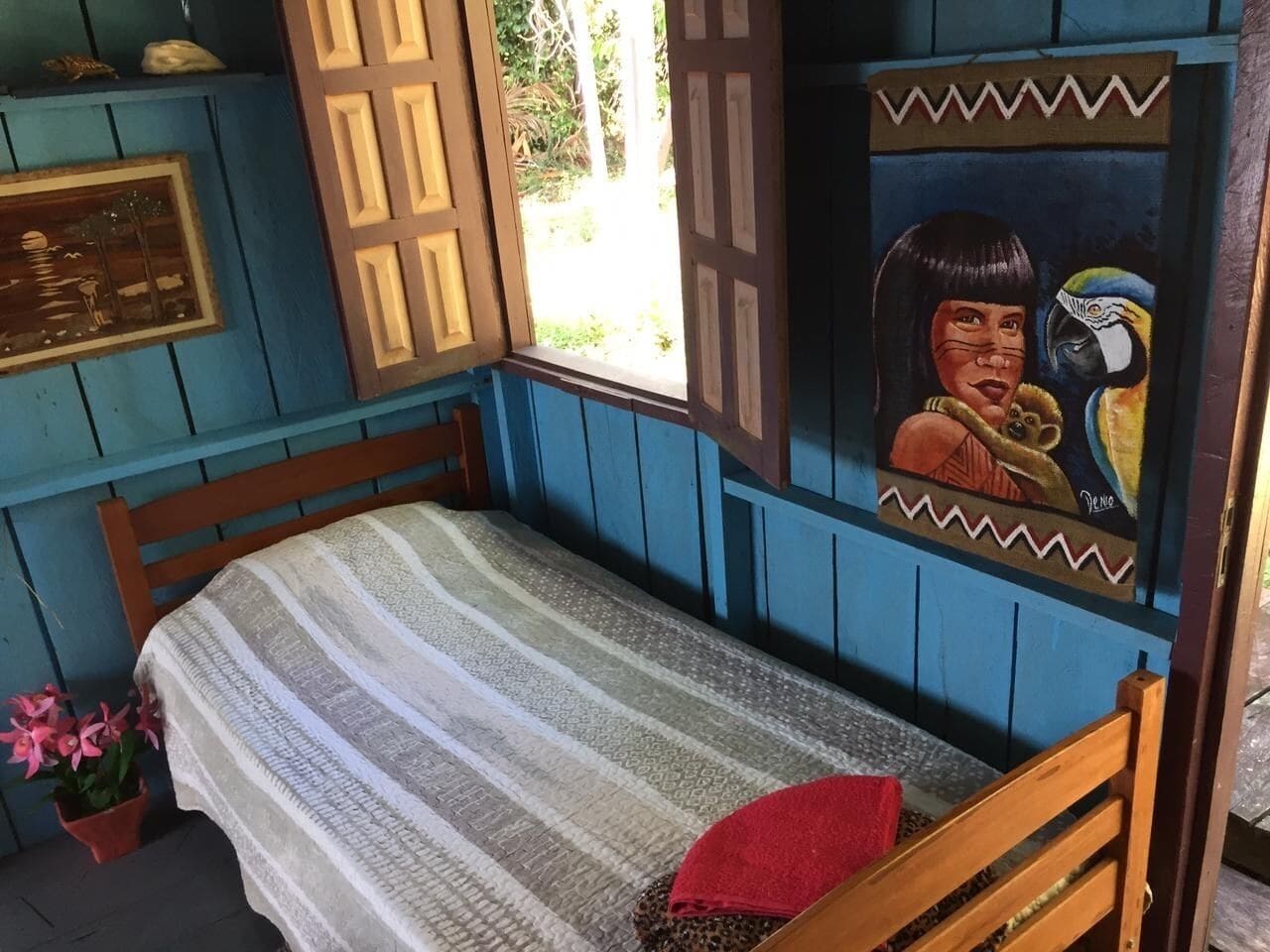 Una cama individual con una colcha a rayas, un almoháron rojo y una ventana con vistas a la vegetación.