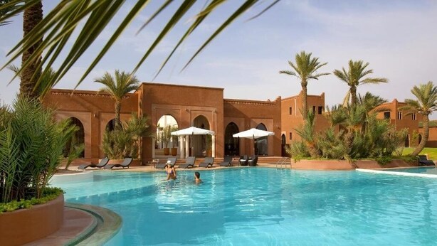 A swimming pool with lounge chairs and umbrellas, surrounded by palm trees and buildings with arches. - Residence Dar Lamia Marrakech : 2 Bedroom Villa with Private Pool (Dar Caïd Layadi)