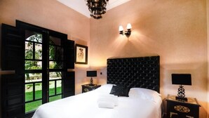 A bedroom with a large bed, two bedside tables, a chandelier, and a window with a view of greenery. - Residence Dar Lamia Marrakech : 2 Bedroom Villa with Private Pool (Dar Caïd Layadi)