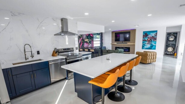 A modern kitchen with a central island, bar stools, a built-in oven, and a large TV above the fireplace.